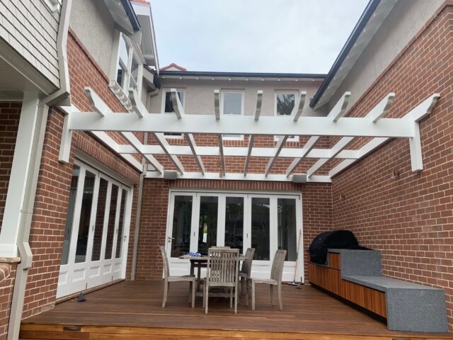 White pergola frames timber deck with dining set and built-in barbecue in brick courtyard.