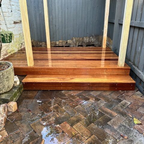 Compact hardwood deck with single step and pergola posts, bordered by grey fences.