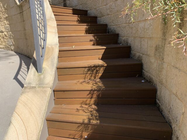 Curved timber outdoor steps with recessed lights, bordered by sandstone retaining wall and metal handrail.
