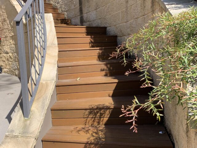 Timber outdoor stairs with curved metal balustrade, sandstone retaining walls and garden shrubs.