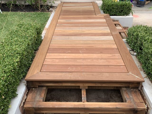 Timber entry decking under construction, boards fitted with subframe exposed at lower steps, hedges alongside.