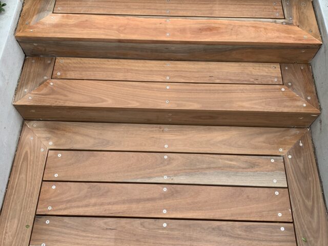 Hardwood decking steps with mitred corners and stainless screws, flanked by concrete planters.