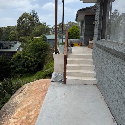 Concrete path and stairs with exposed posts beside grey brick house, sandstone outcrop and bushland.
