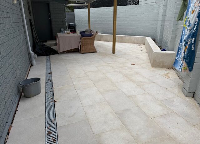 Tiled courtyard with linear trench drain, pergola posts, and low rendered retaining wall.