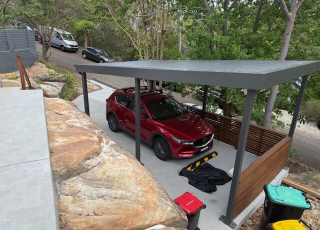 Steel carport with flat roof and timber slat screen on concrete pad, red SUV parked.