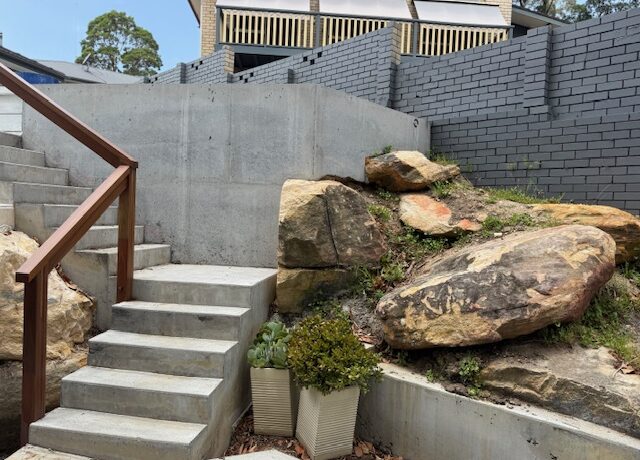 Concrete retaining wall and steps with timber handrail beside sandstone boulders and planters.