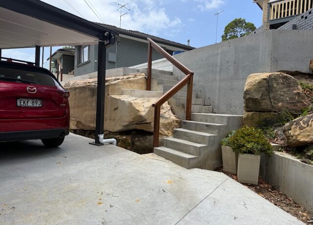 Tiered concrete steps with timber handrails beside sandstone retaining walls and carport driveway.