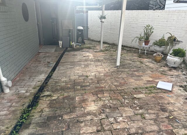 Weathered brick patio with drain and pergola posts, potted plants along white wall.