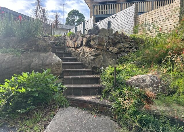 Brick garden steps and stone retaining wall with overgrown plants, leading to raised patio.