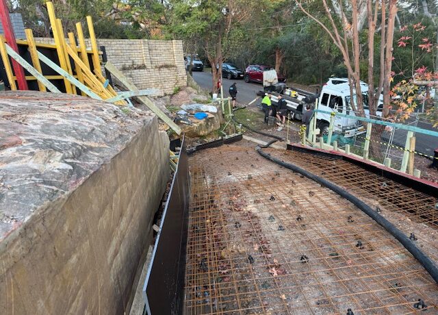 Curved driveway formwork with steel reinforcement on sloping site, workers and trucks nearby.
