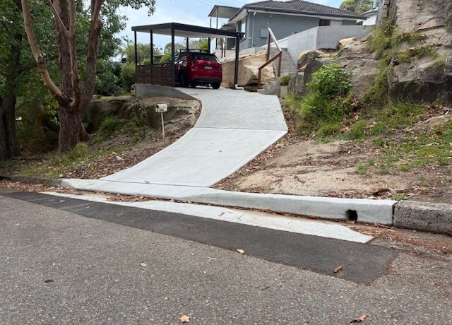 Steep concrete driveway rising from street to carport with red car, sandstone rock slope, and timber handrail.