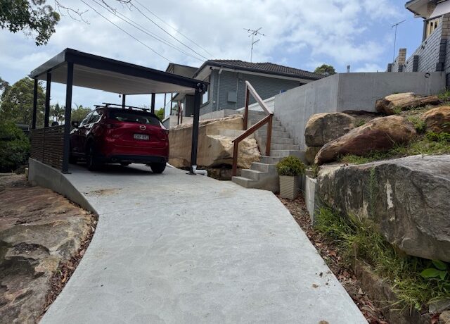 Sloped concrete driveway to carport with red SUV, sandstone retaining walls and stairs with timber handrail.