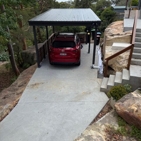Concrete driveway and carport with red SUV, adjacent timber-railed steps and sandstone landscaping.