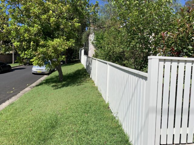 White timber slat boundary fence along suburban verge with green lawn and trees.