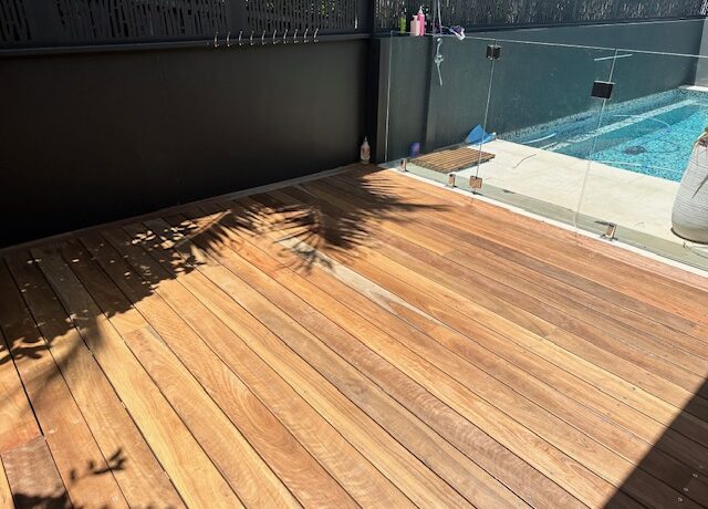 Sunlit timber poolside deck with glass balustrade and black privacy screen.