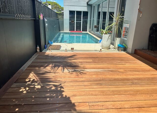 Hardwood poolside deck with glass balustrade outside a modern home.