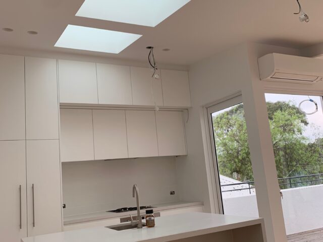 Modern white kitchen under three skylights, island sink, unfinished ceiling lights, opening to deck.