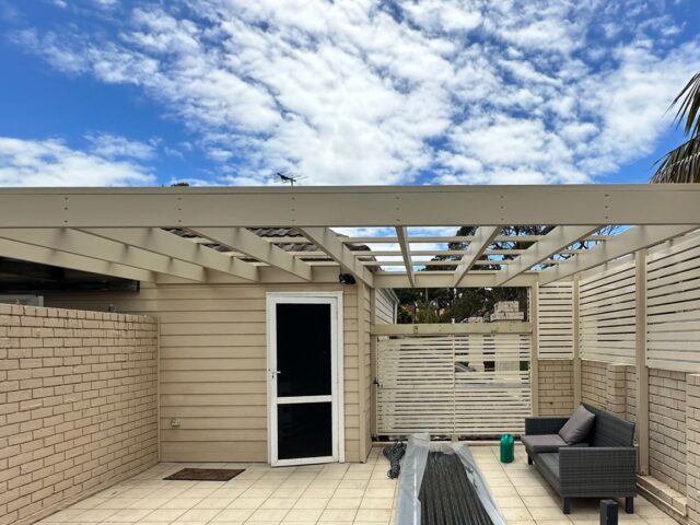 Timber pergola frame under construction over tiled courtyard with slatted privacy screens and outdoor sofa.