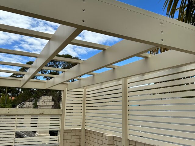 Beige pergola frame with slatted privacy screens, brick patio, and outdoor lounge chair.