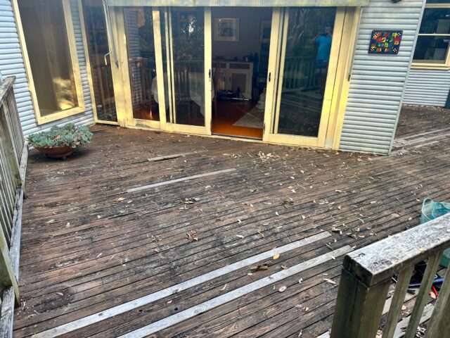 Weathered outdoor timber deck with worn boards and leaves by sliding glass doors.