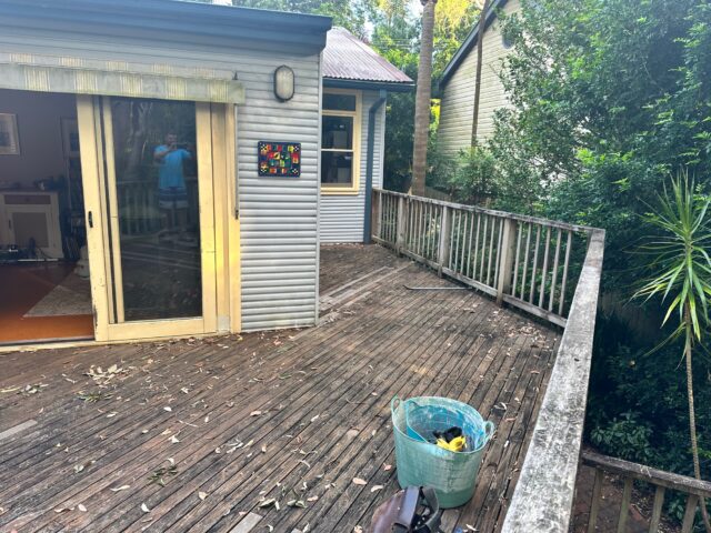 Weathered timber deck with balustrade and sliding door, tools bucket, leafy garden.