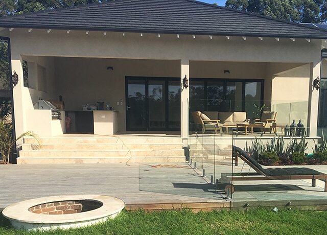 Covered alfresco with outdoor kitchen, deck and glass balustrade, fire pit and lawn.