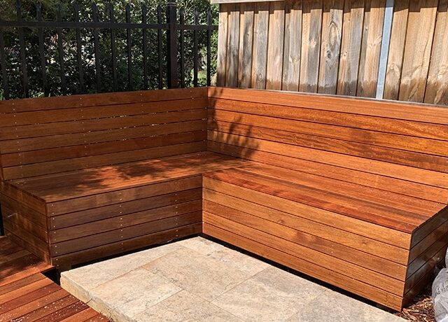 Built-in L-shaped hardwood bench on stone pavers beside timber fence and metal railing.