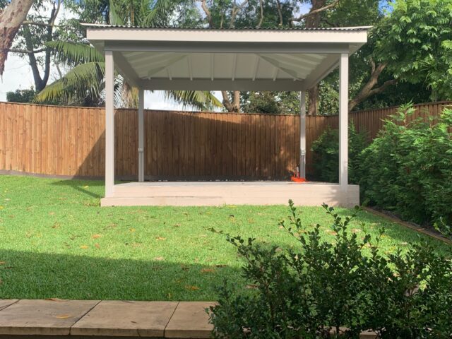 Freestanding pergola with metal roof on concrete slab, landscaped backyard lawn.