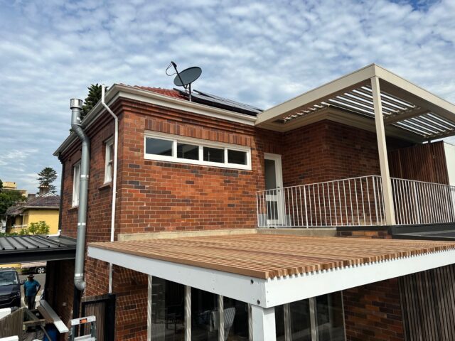 Brick home with upper deck, white balustrade and louvered pergola; timber slat roof below.