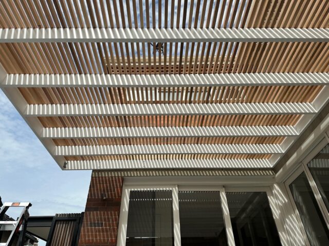 Timber slat pergola on white frame casting striped shade over patio doors.