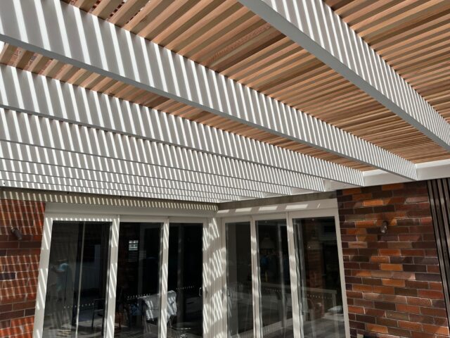 Timber batten pergola on white frame casting striped shade over brick wall and sliding glass doors.