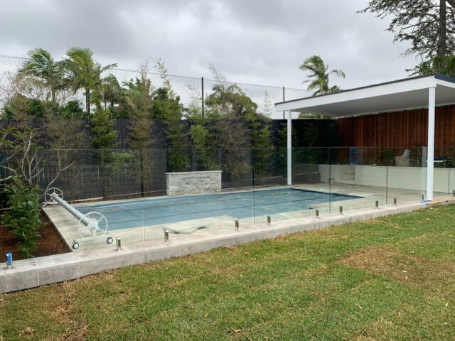 Backyard pool with glass fencing, tiled surround, covered alfresco pergola and tropical landscaping.