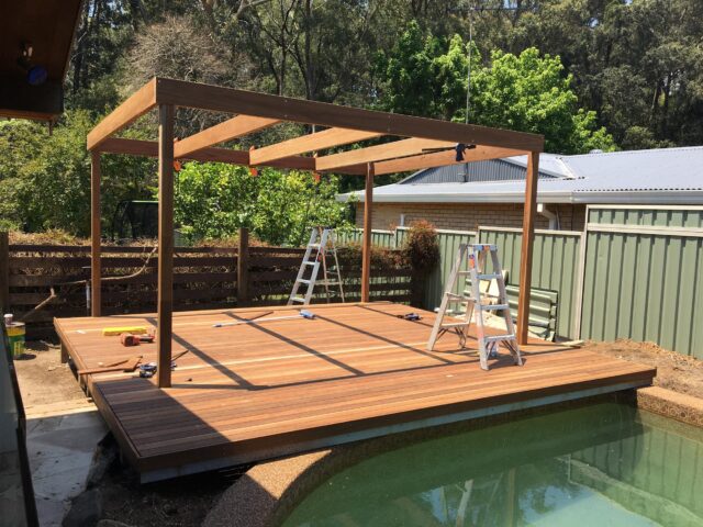 Timber poolside deck with pergola frame under construction, ladders and tools in backyard.