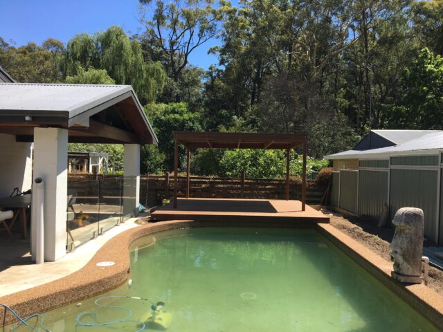 Timber pergola and raised deck beside backyard pool with glass balustrade.