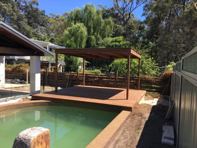 Timber poolside deck with pergola under construction in a leafy backyard.