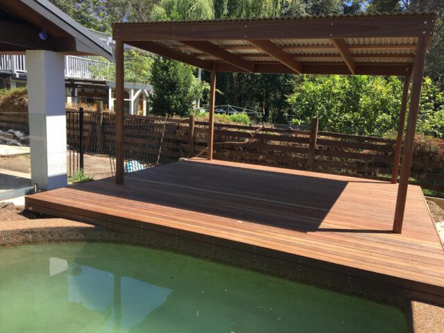 Timber deck with corrugated-roof pergola beside a backyard pool and garden.