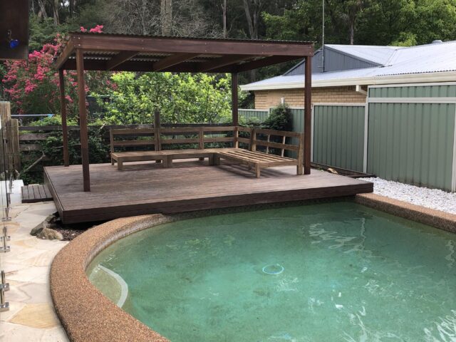 Timber poolside deck with pergola and bench seating beside curved pool.