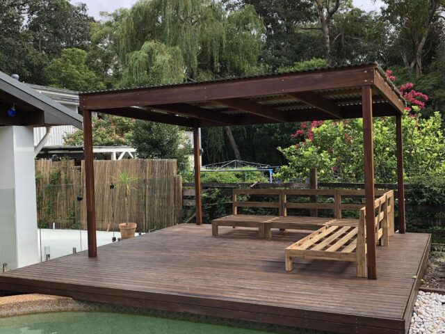 Covered timber deck beside swimming pool, pergola roof and bench seating in leafy yard.