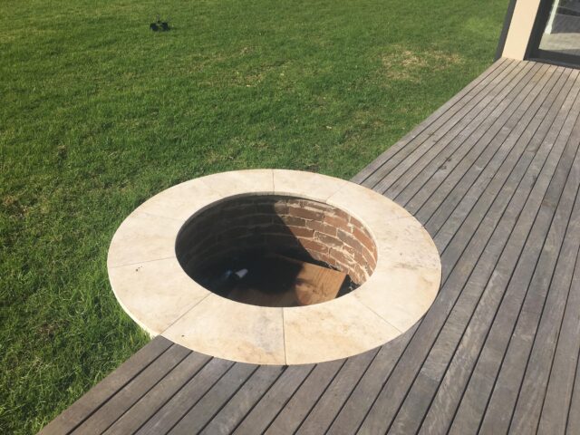 Circular stone-edged brick fire pit recessed into timber deck beside lawn.