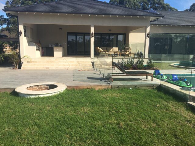 Covered patio with outdoor kitchen, stone steps, timber deck, glass pool fence and lawn fire pit.