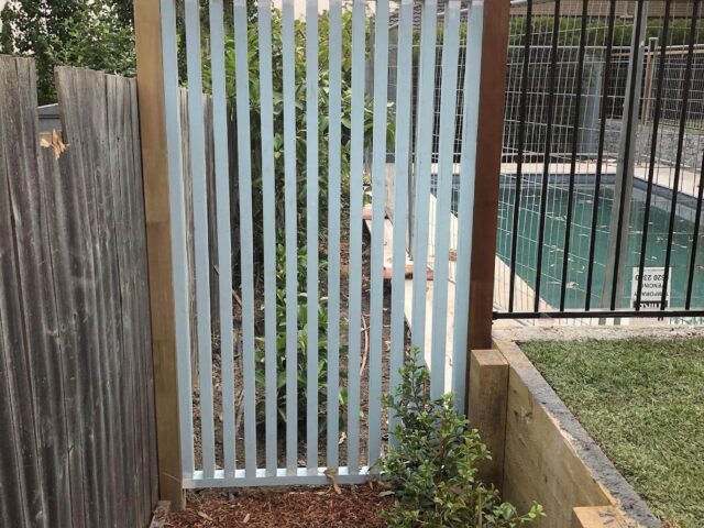 Timber-framed vertical slat gate beside pool fence, with mulched garden bed and shrubs.