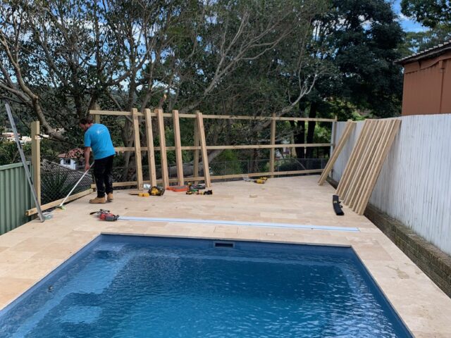 Tradesperson installing timber privacy screen beside pool with light stone pavers, tools nearby.