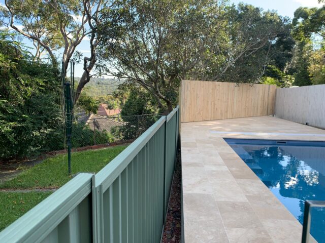 Backyard pool with travertine paving, timber privacy fence, and green metal boundary fence.