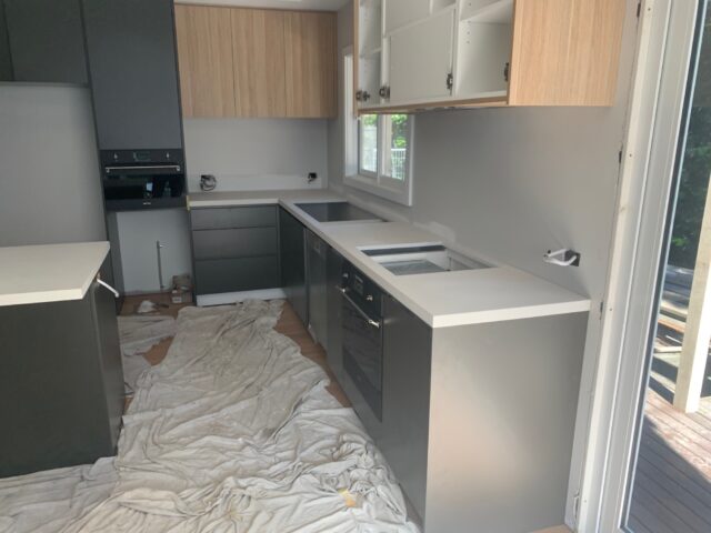 Kitchen renovation underway with grey cabinets, timber uppers, white benchtops, oven installed, drop cloths.
