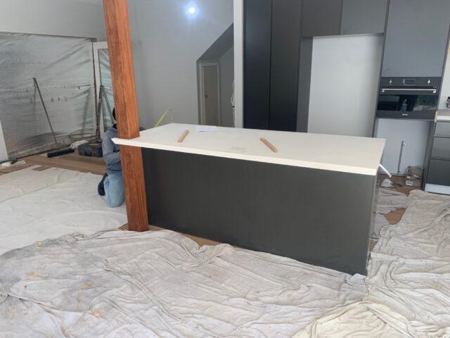 Kitchen under renovation with new island bench, timber support post, grey cabinetry, drop sheets.