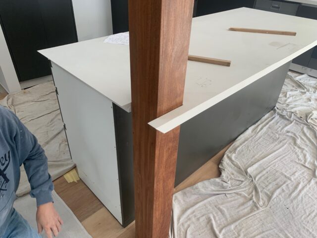 Kitchen renovation: white island benchtop notched around timber post, drop sheets and dark cabinetry.