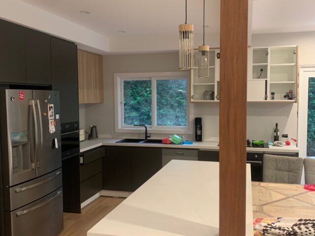 Modern kitchen renovation with white island, timber column, pendant lights, black cabinetry and stainless fridge.