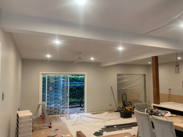 Open-plan living area mid-renovation with sliding doors, recessed lights, timber post, drop sheets and tools.