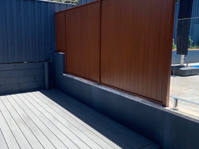 Grey composite deck with step and timber privacy screen beside glass pool fence.