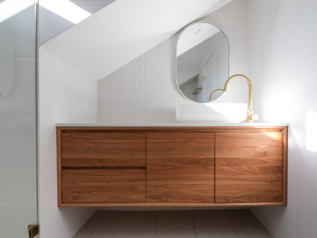 Floating timber vanity, oval mirror, vessel sink and brass tap under sloped ceiling.